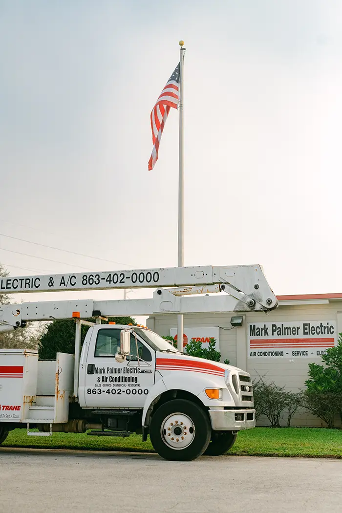 Mark Palmer branded van parked in front of the Mark Palmer office building, with an American flag flying in the background over a clear blue sky.